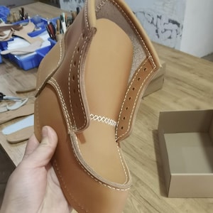 May include: A tan leather shoe upper with white stitching. The shoe is turned inside out, showing the lining and the stitching details.