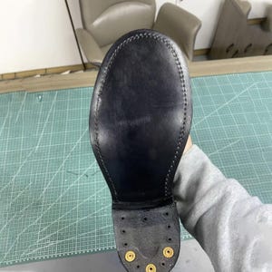 Op de afbeelding: Close-up van een zwarte leren schoenzool, met gedetailleerde stiksels rondom. De zool heeft een gladde, gepolijste afwerking en messingkleurige hardware bij de hiel. De schoenzool is waarschijnlijk voor een laars of nette schoen.