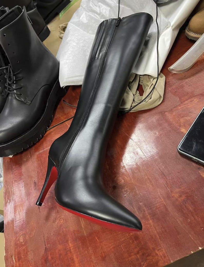 Puede incluir: Bota alta de cuero negro con punta y tac&oacute;n de aguja. El tac&oacute;n es rojo y la suela tambi&eacute;n es roja. La bota tiene una cremallera en el lateral. Otra bota negra es visible en el fondo.