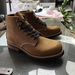 May include: A pair of brown leather boots with dark brown laces. The boots have a rugged, workwear style with a stitched sole and a sturdy, textured outsole. The leather has a matte finish.