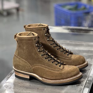 May include: A pair of brown suede work boots with black laces. The boots have a sturdy design with a light-colored sole and a stacked heel. The boots are designed for durability and are suitable for various outdoor activities.