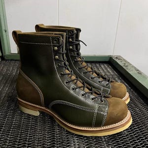 Puede incluir: Un par de botas de trabajo de cuero verde oliva y marrón. Las botas tienen una puntera redondeada, un diseño con cordones y costuras blancas. Las suelas son de color marrón claro.