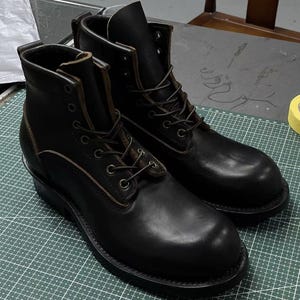Op de afbeelding: Een paar zwarte leren werklaarzen met vetersluiting. De laarzen hebben een ronde neus, een stevige zool en metalen vetergaten. De stiksels zijn zichtbaar en de laarzen lijken versleten.