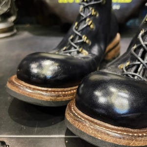 May include: Close-up of a pair of black leather boots with a rounded toe and lace-up design. The boots have a thick, brown sole and gold-colored eyelets. The boots appear to be new and unworn.