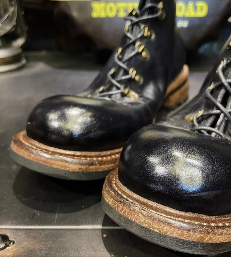 May include: Close-up of a pair of black leather boots with a rounded toe and lace-up design. The boots have a thick, brown sole and gold-colored eyelets. The boots appear to be new and unworn.