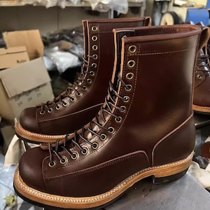 Puede incluir: Un par de botas de trabajo de cuero marrón con diseño de cordones. Las botas tienen múltiples ojales, una suela resistente y una puntera reforzada. El cuero tiene un color marrón oscuro intenso y las botas parecen nuevas.