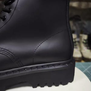 May include: Close-up of a black combat boot. The boot features a smooth, matte finish, a thick lug sole, and black laces. The stitching is visible along the seams. The boot is a classic style, suitable for various outfits.