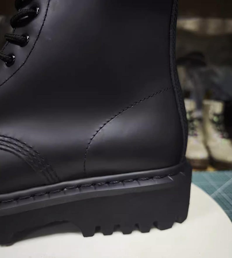 May include: Close-up of a black combat boot. The boot features a smooth, matte finish, a thick lug sole, and black laces. The stitching is visible along the seams. The boot is a classic style, suitable for various outfits.