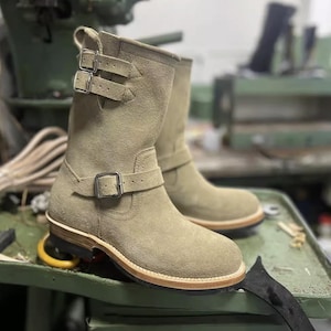 May include: Olive green suede boots with buckle fastenings. The boots are mid-calf length, with a rounded toe and a leather sole. The boots are displayed on a green surface.