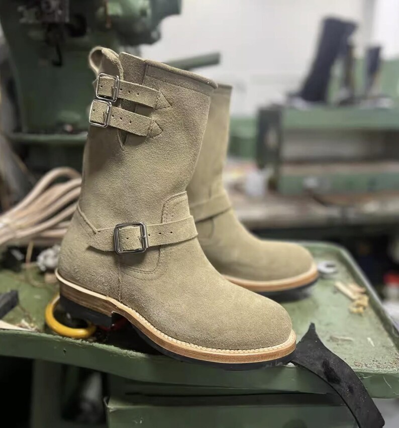 May include: Olive green suede boots with buckle fastenings. The boots are mid-calf length, with a rounded toe and a leather sole. The boots are displayed on a green surface.