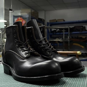 Op de afbeelding: Een paar zwarte leren werklaarzen met een vetersluiting. De laarzen hebben een ronde neus, een stevige zool en een middelhoge schacht. De laarzen zijn ongeveer 20 cm hoog.