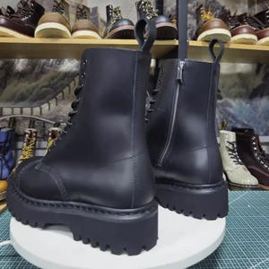 May include: A pair of black leather combat boots with a chunky, lugged sole. The boots have a lace-up front and a zipper on the back. The boots are displayed on a white surface, with other boots visible in the background.