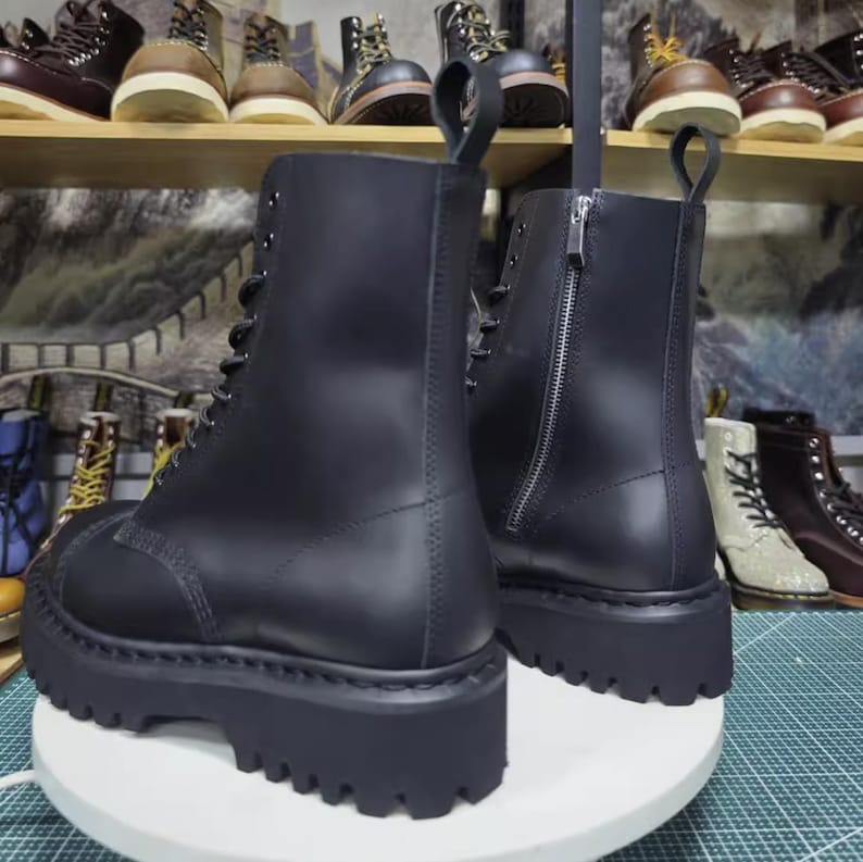 May include: A pair of black leather combat boots with a chunky, lugged sole. The boots have a lace-up front and a zipper on the back. The boots are displayed on a white surface, with other boots visible in the background.