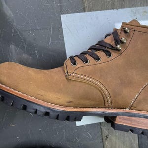 May include: A brown leather work boot with a rugged design. The boot features a lace-up closure, metal eyelets, and a thick black rubber sole. The stitching is visible, and the boot has a worn, textured appearance. The sole measures approximately 1 inch in height.
