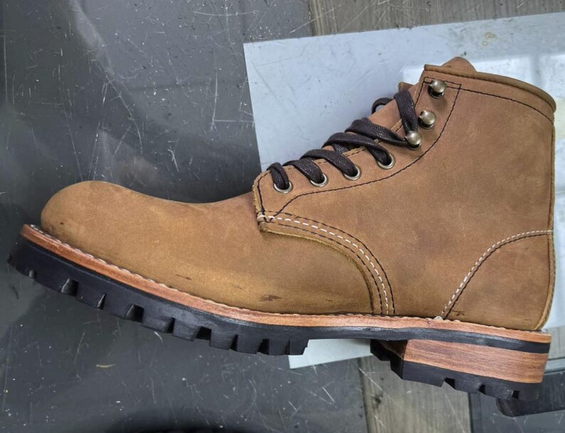 May include: A brown leather work boot with a rugged design. The boot features a lace-up closure, metal eyelets, and a thick black rubber sole. The stitching is visible, and the boot has a worn, textured appearance. The sole measures approximately 1 inch in height.