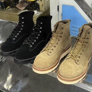 Könnte beinhalten: Vier Wildleder-Arbeitsstiefel in Schwarz und Beige. Die schwarzen Stiefel haben weiße Nähte und silberne Ösen. Die beigefarbenen Stiefel haben gelbe Schnürsenkel und weiße Sohlen. Die Stiefel werden auf einer Metalloberfläche präsentiert.