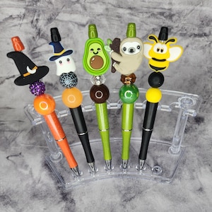 May include: Six colorful pens with different character toppers, including a ghost, a sloth, a bee, an avocado, and a witch. The pens are displayed in a clear plastic stand.