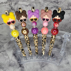 May include: Five beaded pens with leopard print barrels and cartoon girl toppers wearing heart-shaped sunglasses. The pens have different colored beads and toppers in yellow, brown, purple, pink, and black.
