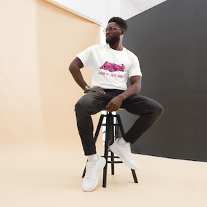 May include: White t-shirt with a pink cartoon creature and the text "WHAT IF I JUST DIDN'T". The person is wearing black jeans and white trainers, sitting on a black stool. The background is beige and black.
