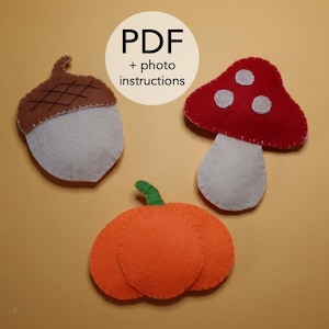 Puede incluir: Decoraciones de otoño de fieltro que incluyen una bellota, una seta y una calabaza. La bellota tiene una tapa marrón y una base blanca. La seta es roja con puntos blancos. La calabaza es naranja. El texto en la imagen dice "PDF + photo instructions."