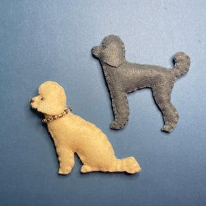 Puede incluir: Dos adornos de fieltro con forma de perro caniche. Uno es amarillo claro con un collar de cuentas, el otro es gris. Ambos están cosidos por los bordes. Los perros están colocados sobre un fondo azul.