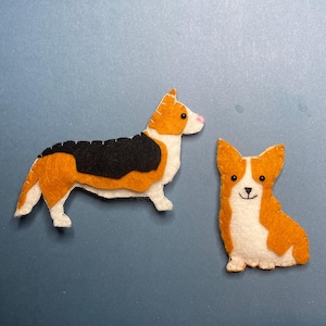 Può includere: Due figure di cane corgi in feltro. Uno è in piedi, con la schiena nera, segni arancioni e bianchi e un naso rosa. L'altro è seduto, con segni arancioni e bianchi e una faccia sorridente. Lo sfondo è blu.
