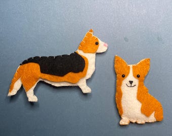 May include: Two felt corgi dog figures. One is standing, with a black back, orange and white markings, and a pink nose. The other is sitting, with orange and white markings, and a smiling face. The background is a solid blue.