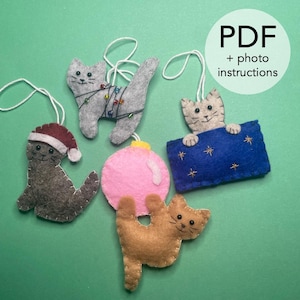 May include: A selection of handmade felt cat decorations in different colours and designs. One grey cat is wrapped in fairy lights, another wears a Santa hat, and a third holds a blue banner with stars. The image includes the text "PDF + photo instructions."