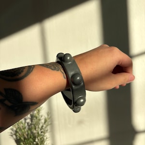 May include: A dark gray bracelet with a series of rounded, raised accents. The bracelet is worn on a wrist, and the design is simple yet striking. The bracelet's color and texture create a modern, minimalist aesthetic.