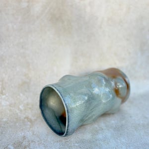 May include: A handmade ceramic tumbler with an organic shape. The exterior has a mottled, light blue-green glaze with brown accents near the rim. The interior is a darker, glossy blue.