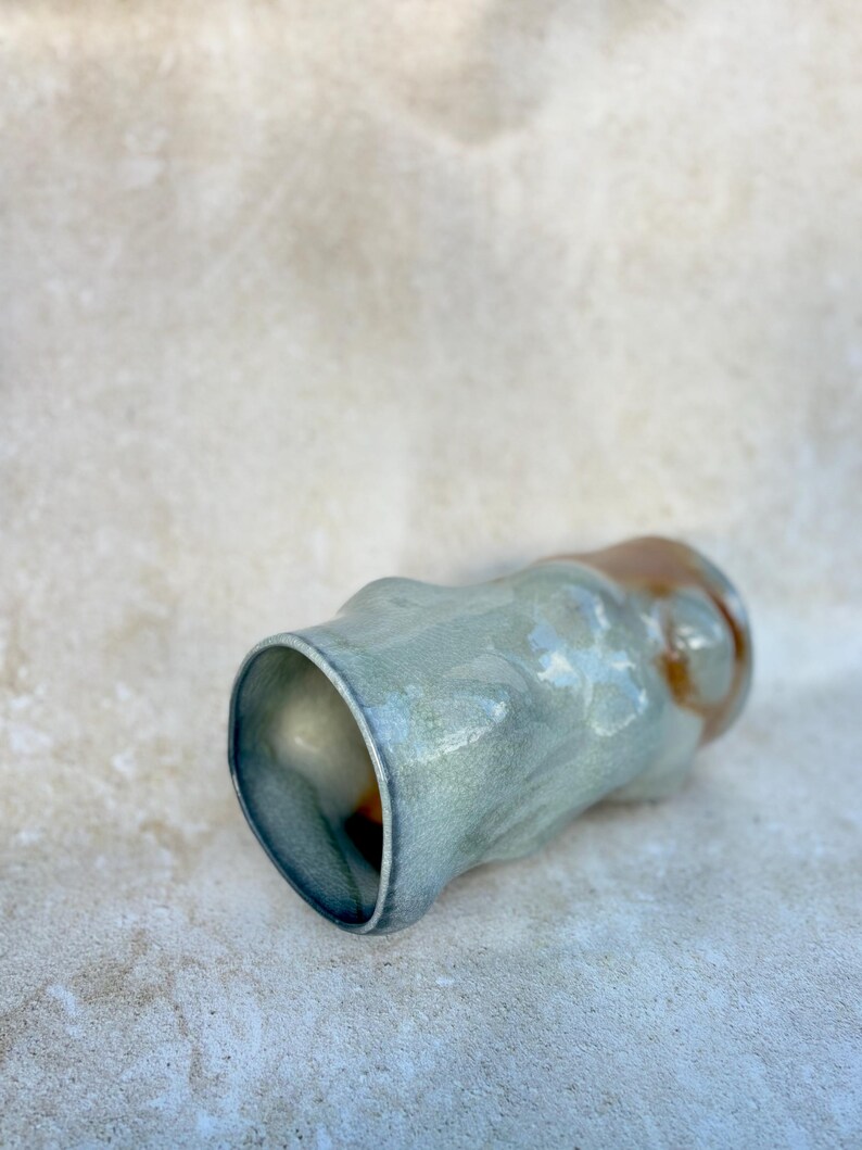 May include: A handmade ceramic tumbler with an organic shape. The exterior has a mottled, light blue-green glaze with brown accents near the rim. The interior is a darker, glossy blue.