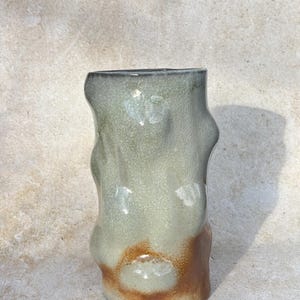 May include: A handmade ceramic vase with an unusual, organic form. The vase has a mix of colours, including light green, brown, and grey. The textured surface enhances its artistic quality. This decorative item is suitable for home decor.