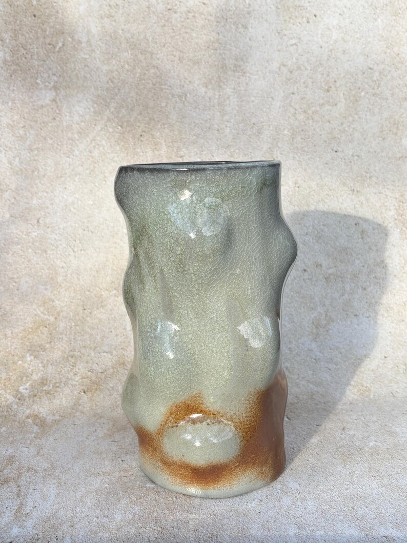 May include: A handmade ceramic vase with an unusual, organic form. The vase has a mix of colours, including light green, brown, and grey. The textured surface enhances its artistic quality. This decorative item is suitable for home decor.