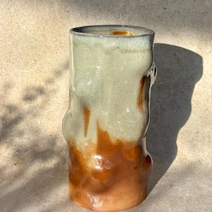 May include: A handcrafted ceramic vase with an unusual, organic form. The vase displays a colour gradient, shifting from light green at the top to a warm, earthy brown at the base. The surface has a crackled texture.