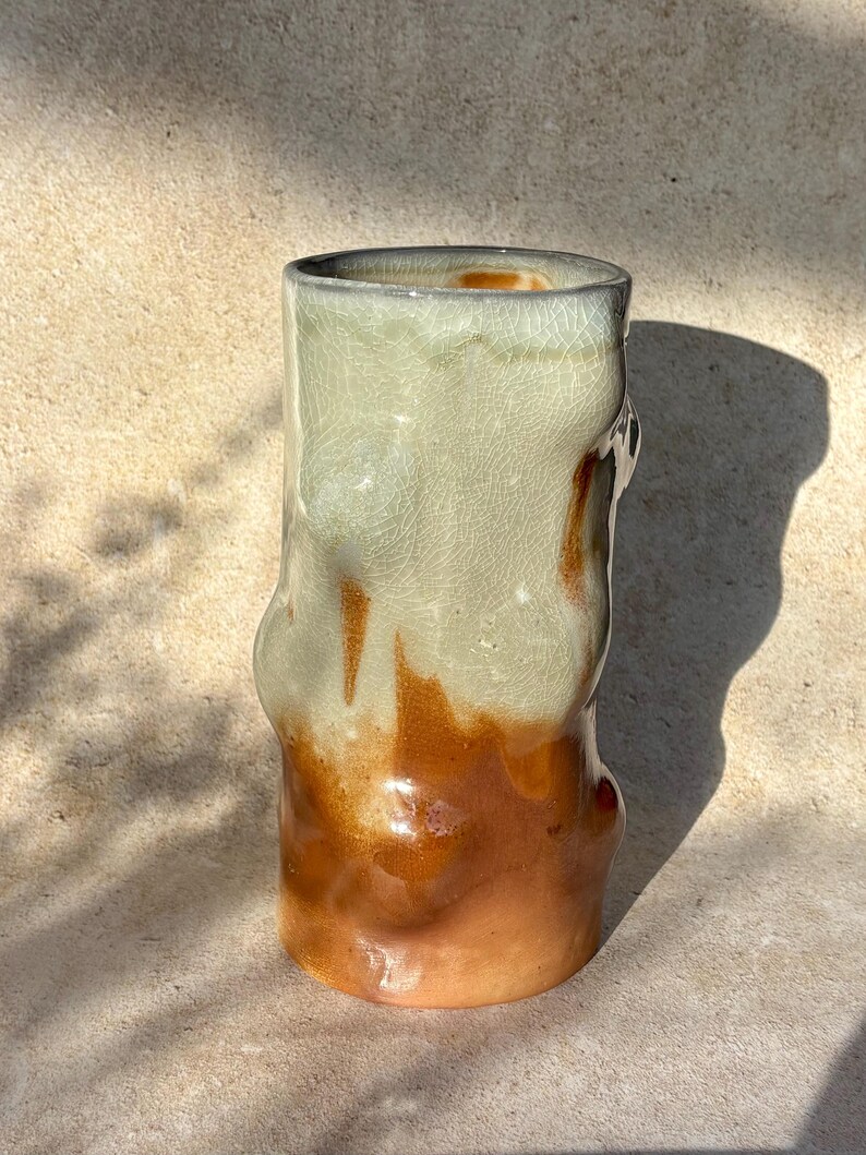 May include: A handcrafted ceramic vase with an unusual, organic form. The vase displays a colour gradient, shifting from light green at the top to a warm, earthy brown at the base. The surface has a crackled texture.