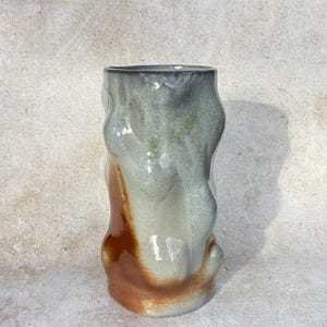 May include: A handcrafted ceramic vase with an unusual, flowing form. The vase has a mix of grey, green, and brown hues, with a shiny surface. The design is abstract and contemporary, ideal for interior decoration.