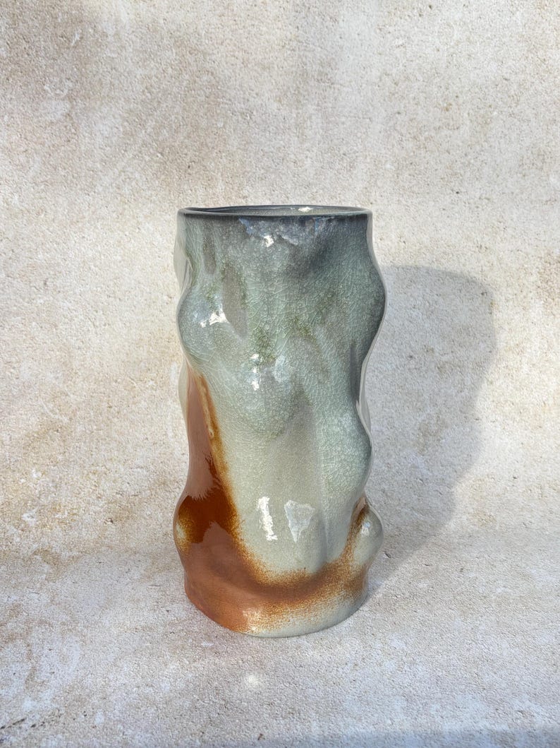 May include: A handcrafted ceramic vase with an unusual, flowing form. The vase has a mix of grey, green, and brown hues, with a shiny surface. The design is abstract and contemporary, ideal for interior decoration.