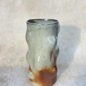 May include: A handmade ceramic vase with an unusual, organic form. The vase has a light green upper section that fades into a brown base. The surface has a crackled texture, enhancing its handcrafted look. The vase is approximately 15 cm tall.