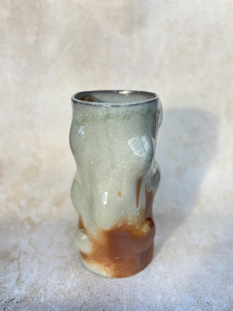 May include: A handmade ceramic vase with an unusual, organic form. The vase has a light green upper section that fades into a brown base. The surface has a crackled texture, enhancing its handcrafted look. The vase is approximately 15 cm tall.