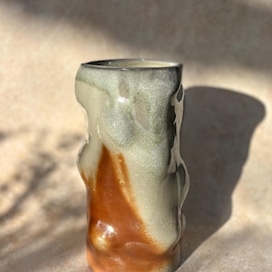 May include: A handmade ceramic vase with a unique, organic shape. The vase features a blend of colors, including a warm, earthy orange at the base that transitions to a creamy white and green at the top. The surface has a crackle glaze.