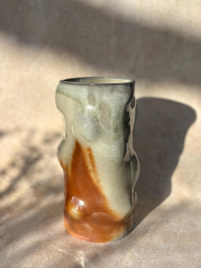 May include: A handcrafted ceramic vase with an unusual, organic form. The vase displays a mix of colours, with a warm, earthy orange at the base that fades into a creamy white and green at the top. The surface has a crackle glaze.