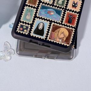Puede incluir: Una funda de tel&eacute;fono negra con un collage de coloridos dise&ntilde;os de sellos postales. Los sellos representan varios personajes y escenas de anime, incluyendo un gato negro, una ni&ntilde;a con un vestido azul y un personaje con pelo largo y rubio. La funda es de pl&aacute;stico resistente.