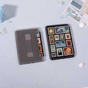 Puede incluir: Una funda de tel&eacute;fono negra con un dise&ntilde;o de sellos coloridos con varios personajes de dibujos animados. La funda est&aacute; abierta, revelando un interior gris y una cubierta protectora transparente. La funda est&aacute; sobre una superficie azul claro.