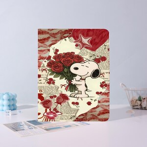 Op de afbeelding: Notitieboek met Snoopy die rode rozen vasthoudt. Het ontwerp bevat een hart, een ster en rozenillustraties, tegen een collage van tekst en lippenstiftvlekken. Romantisch thema.