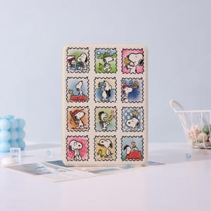 May include: A rectangular beige case featuring a grid of Snoopy illustrations in postage stamp designs. Each stamp showcases Snoopy in various poses, with colorful backgrounds. The case is on a white surface against a light blue backdrop.