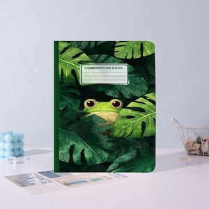 Op de afbeelding: Een compositieboek met een groene kaft met een kikker die door groene bladeren gluurt. De kikker heeft grote, ronde ogen en is omgeven door weelderig gebladerte. De tekst "COMPOSITION BOOK" staat op de kaft.