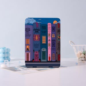 Op de afbeelding: Een decoratief item in de vorm van een boek, met een grillige illustratie van een straat met kleurrijke gebouwen die op boekruggen lijken. De ruggen tonen auteursnamen en boektitels, tegen een achtergrond van een nachtelijke hemel en een halve maan.
