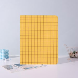 May include: A yellow notebook with a grid pattern in a reddish-brown color. The notebook is rectangular and stands upright on a white surface. A small printed card is in front of the notebook.