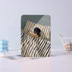 Peut inclure: Un chien teckel noir et marron lisant un livre au lit. Le lit a une couette et un oreiller à rayures. Le chien est au centre de l'image, sur fond bleu clair.