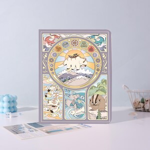 May include: A rectangular notebook featuring a stylized illustration of Appa flying over mountains within a circular frame, surrounded by elemental symbols and smaller panels depicting creatures like a badger-mole and fish in a muted color palette.